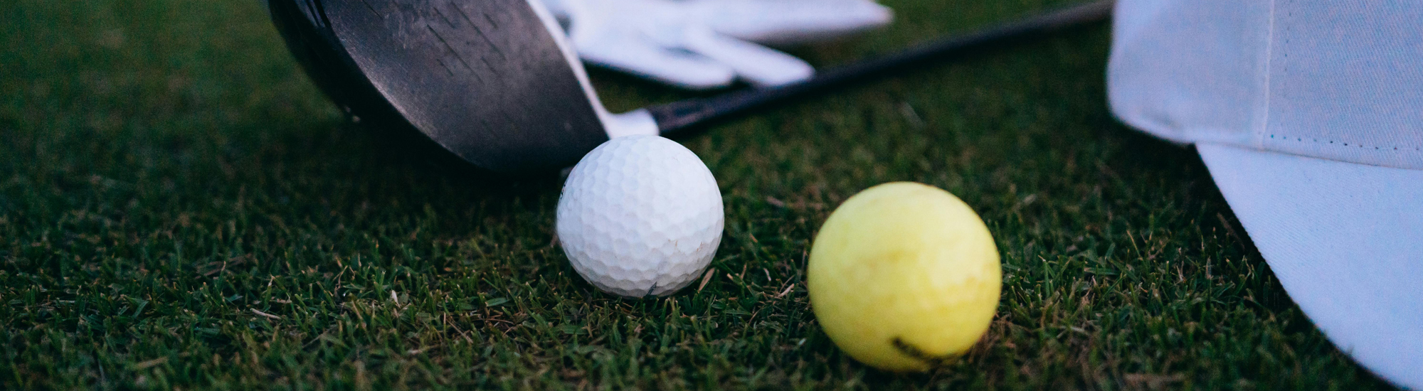 A golf club next to a white golf ball and a yellow ball on green grass, with a white cap partially visible.
