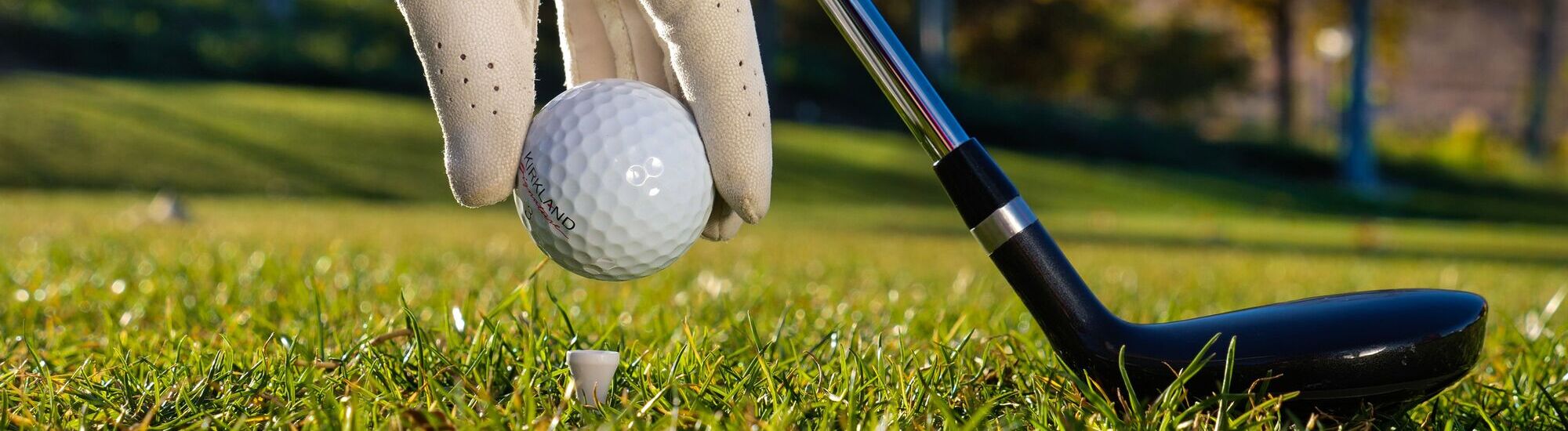 A gloved hand holding a golf ball above a tee on a grassy field, with a golf club nearby.