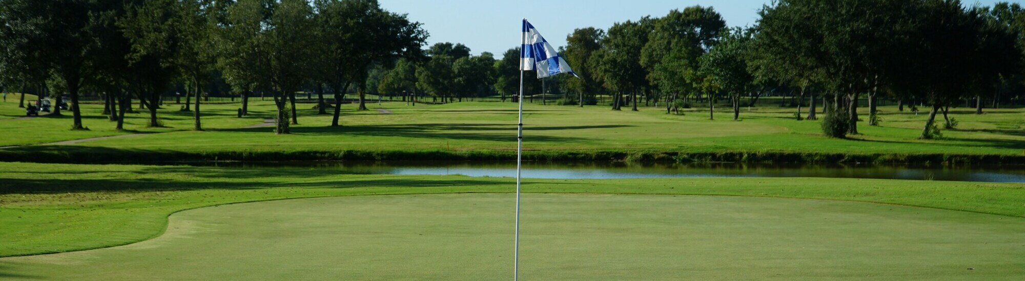 A golf course with a flag on the green, surrounded by trees and a pond in the background.