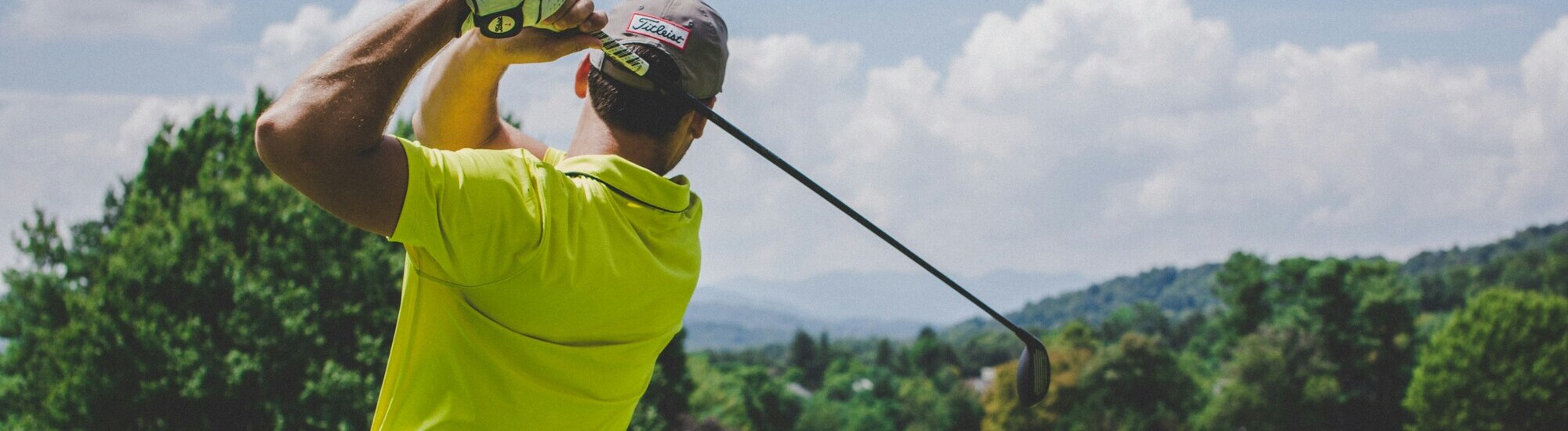 A golfer in a bright yellow shirt swings a club against a backdrop of trees and mountains under a cloudy sky.