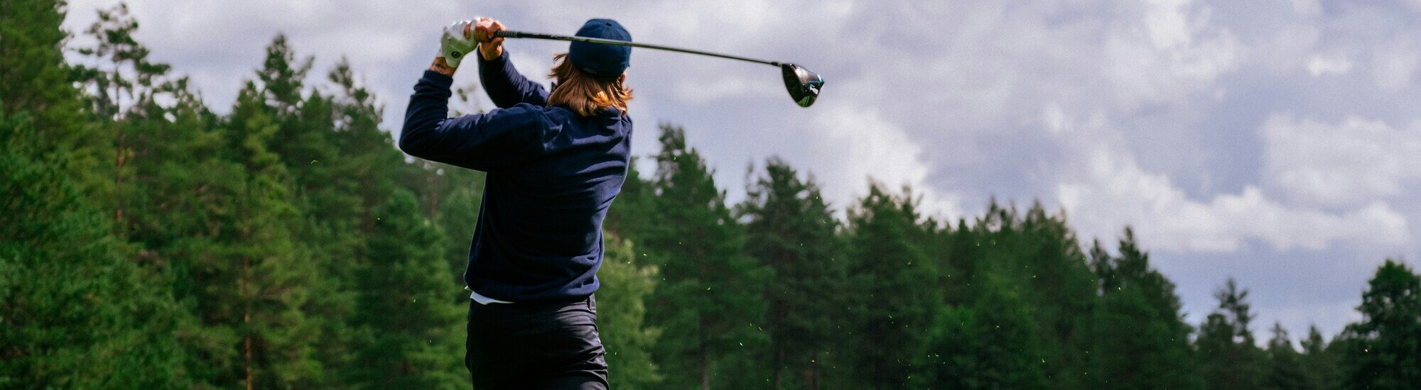A golfer swings a driver on a lush green course under a cloudy sky, surrounded by trees.
